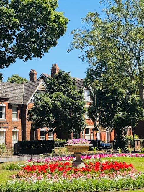 A well-maintained front garden in Hale Village featuring a large, ornate stone planter filled with pink flowers positioned centrally amidst a vibrant flower bed with red and pink blooms. The garden is