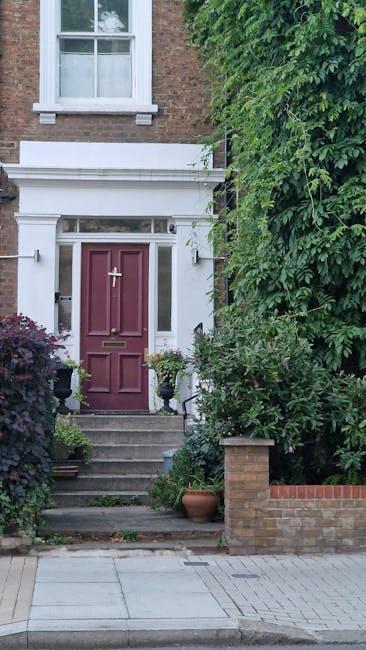 A well-maintained front garden in Hale Village featuring a large, ornate stone planter filled with pink flowers positioned centrally amidst a vibrant flower bed with red and pink blooms. The garden is
